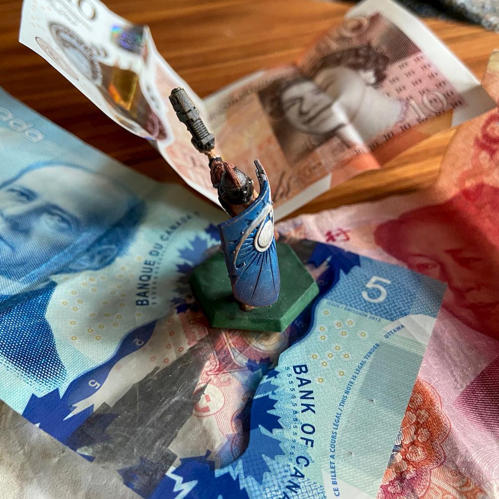 A gaming miniature with a shield and a gun, standing on Canadian, Chinese, and British banknotes