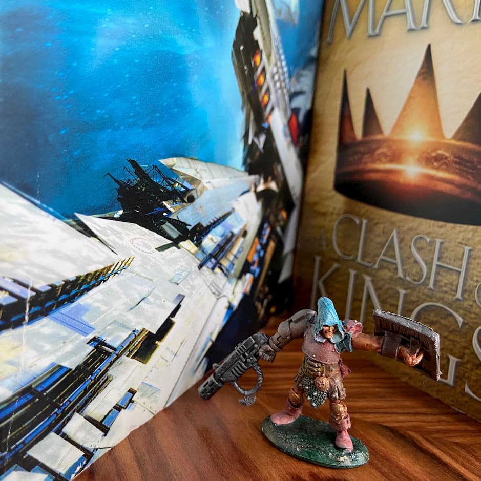 A gaming miniature standing between two books, one fantasy and one sci-fi