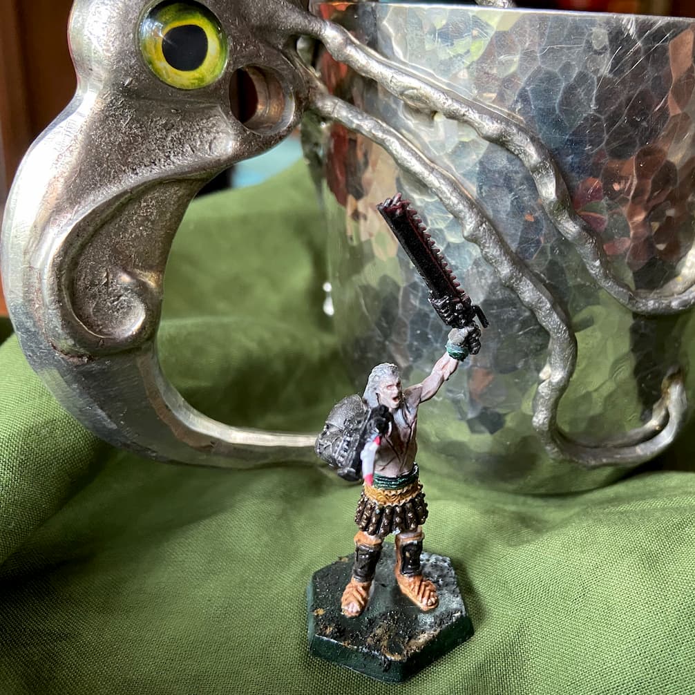 A miniature figure waving a gun and a chainsword, in front of a pewter goblet with a squid shaped handle