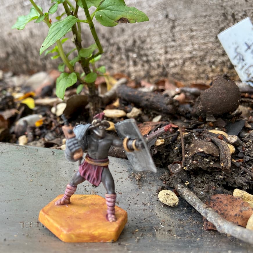 A miniature of a Warhammer 40k cultist, standing on a trowel, in front of a mint plant